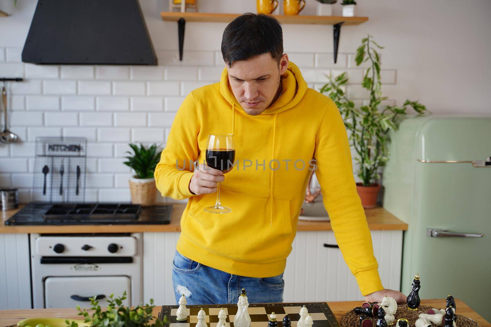 Young man playing chess on kitchen table and drinking red wine. Male with alcohol plays in logical board game with himself, standing in kitchen