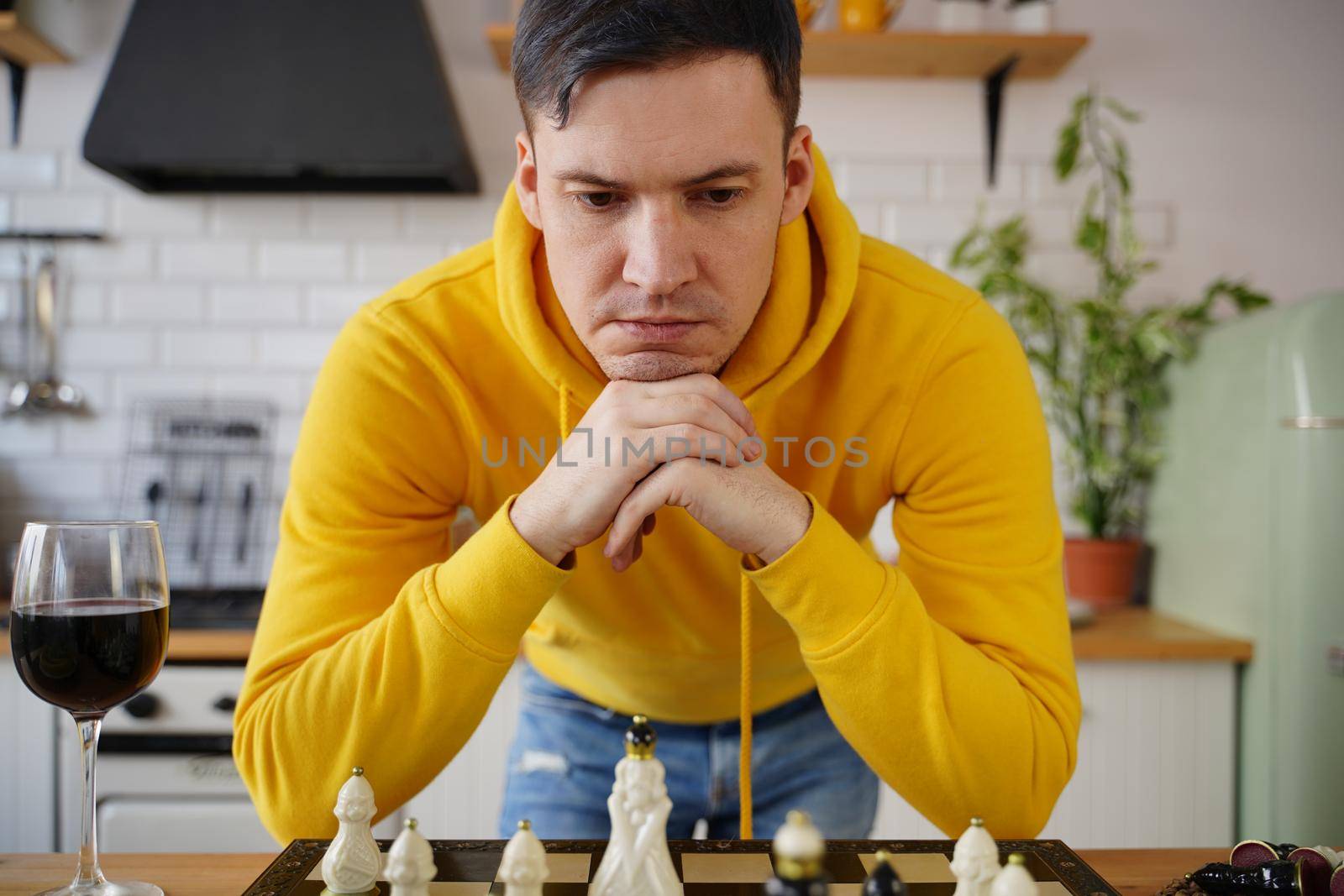 Young man playing chess on kitchen table. Male plays in logical board game with himself, standing in kitchen