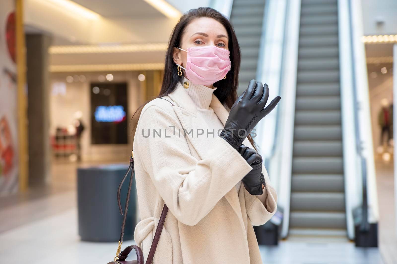 beautiful woman in pink medical mask putting on leather gloves in a public place on background escalator