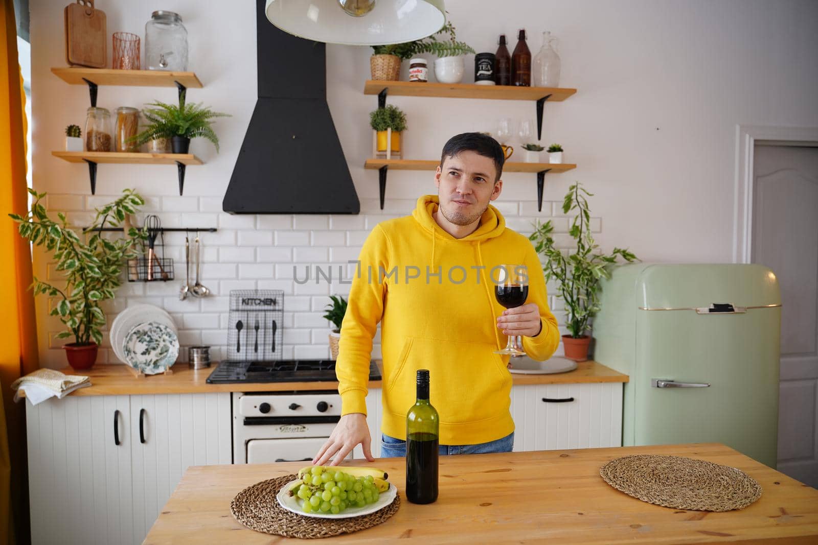 Relaxed young man poses with glass of red wine, standing at kitchen table. Adult guy resting with alcohol and fruits in kitchen