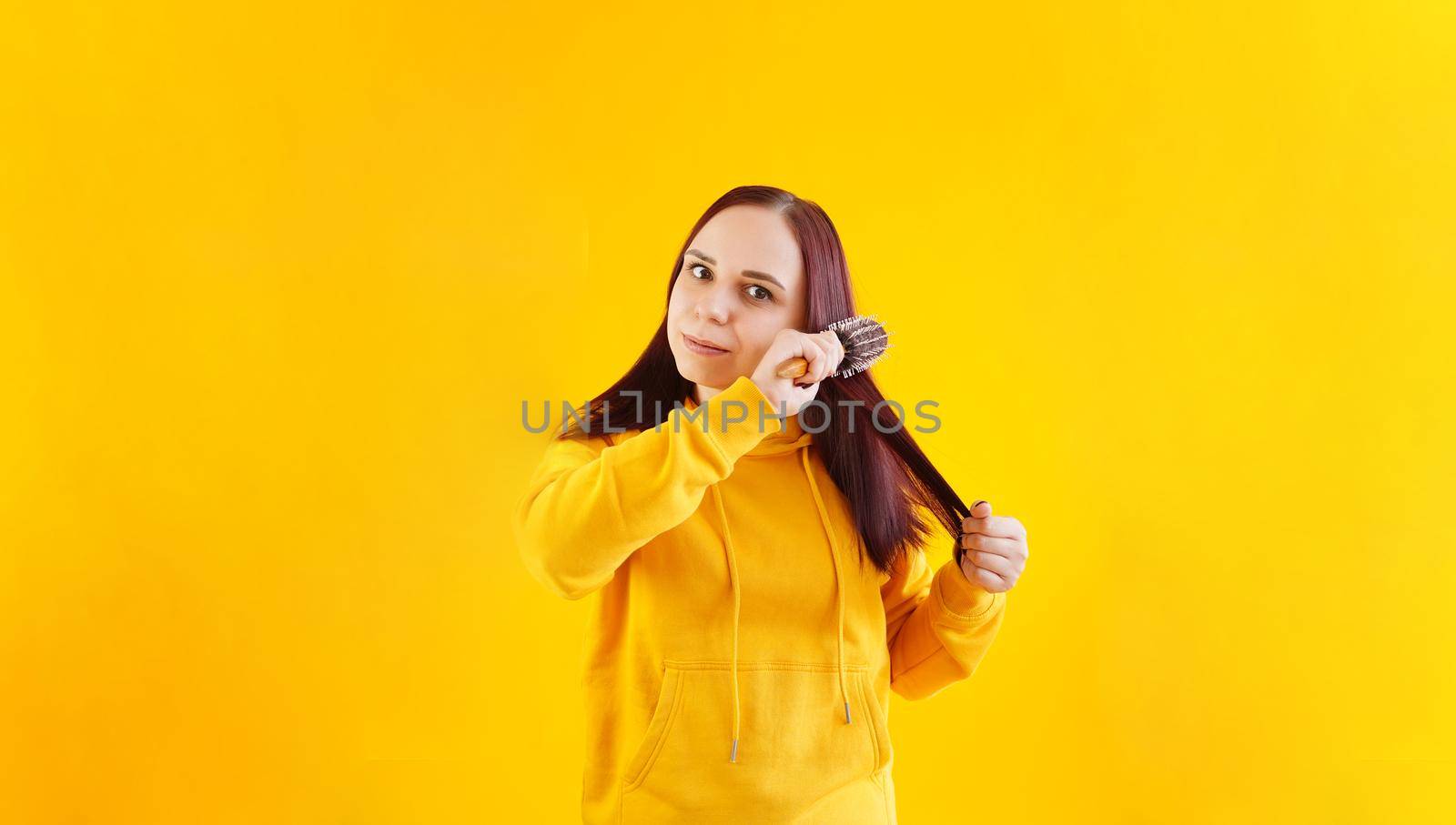Portrait of young woman combing her hair on yellow background. Brunette in yellow hoodie preens, while looking at camera