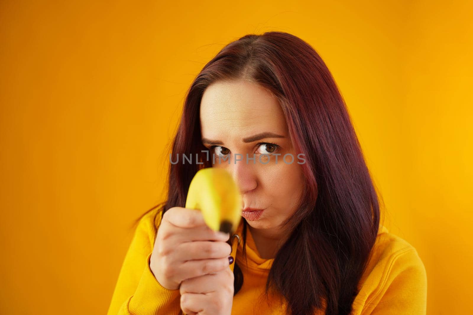 Portrait of young woman with banana on yellow background. Close up of female in yellow hoodie plays with fruit, imagining it as weapon