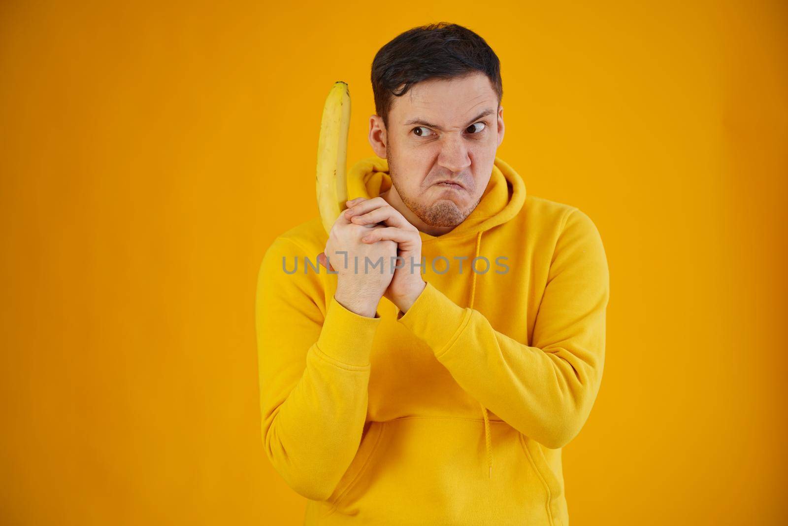 Portrait of young man with banana on yellow background. Close up of handsome guy in yellow hoodie plays with fruit, imagining it as weapon