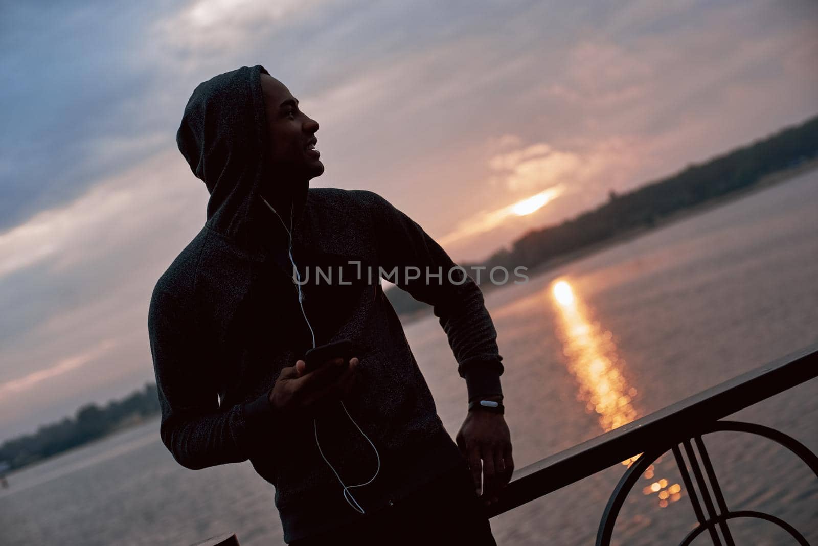 Close-up photo of muscular african male model in hood. He have a little rest after running while is listening to music. Meeting beautiful sunrise