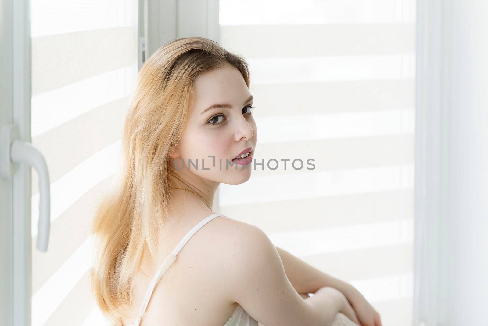 beautiful young woman sits by the window in a white room with her back half-turned.