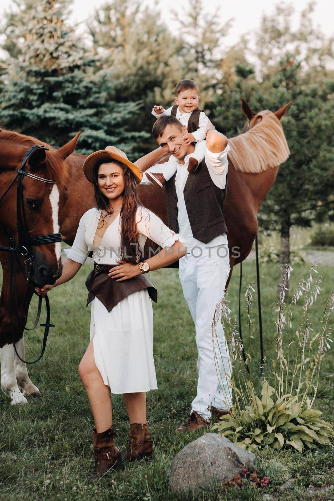 A family in white clothes with their son stand near two beautiful horses in nature. A stylish couple with a child are photographed with horses.