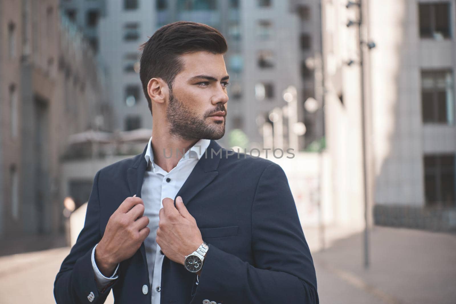 Confident businessman. Confident young man in full suit adjusting his sleeve and looking away while standing outdoors with cityscape in the background by friendsstock