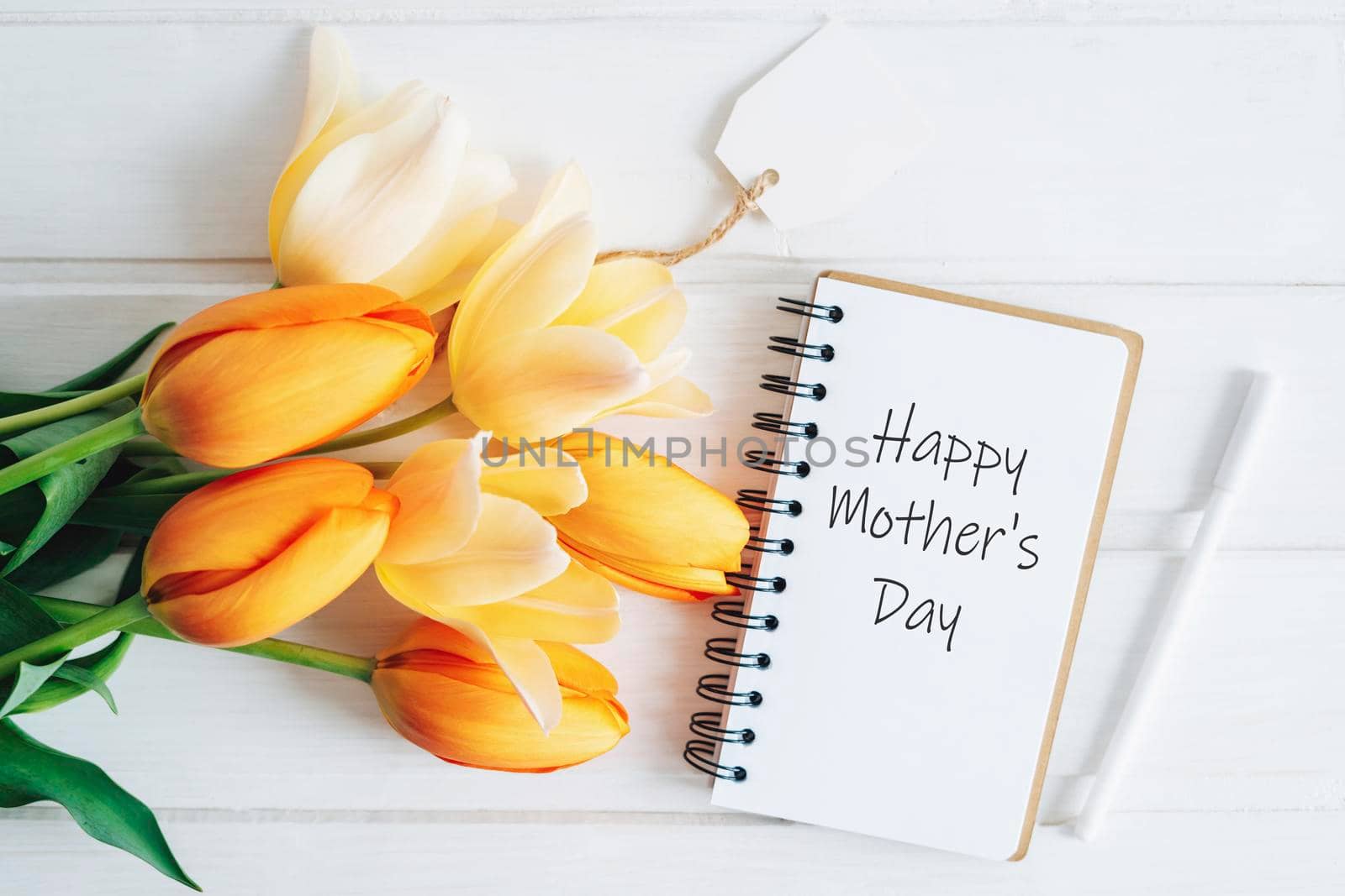 The notebook says Happy Mother's Day. A notebook, a pen and a bouquet of tulips lie on a wooden table. A greeting card.