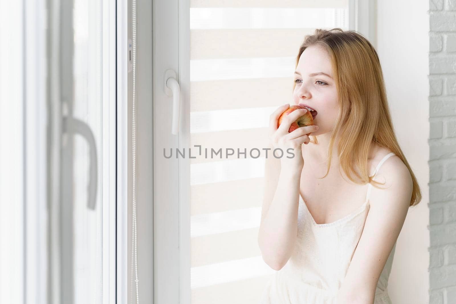 beautiful teen girl in a white dress eats an apple by the window in a bright room