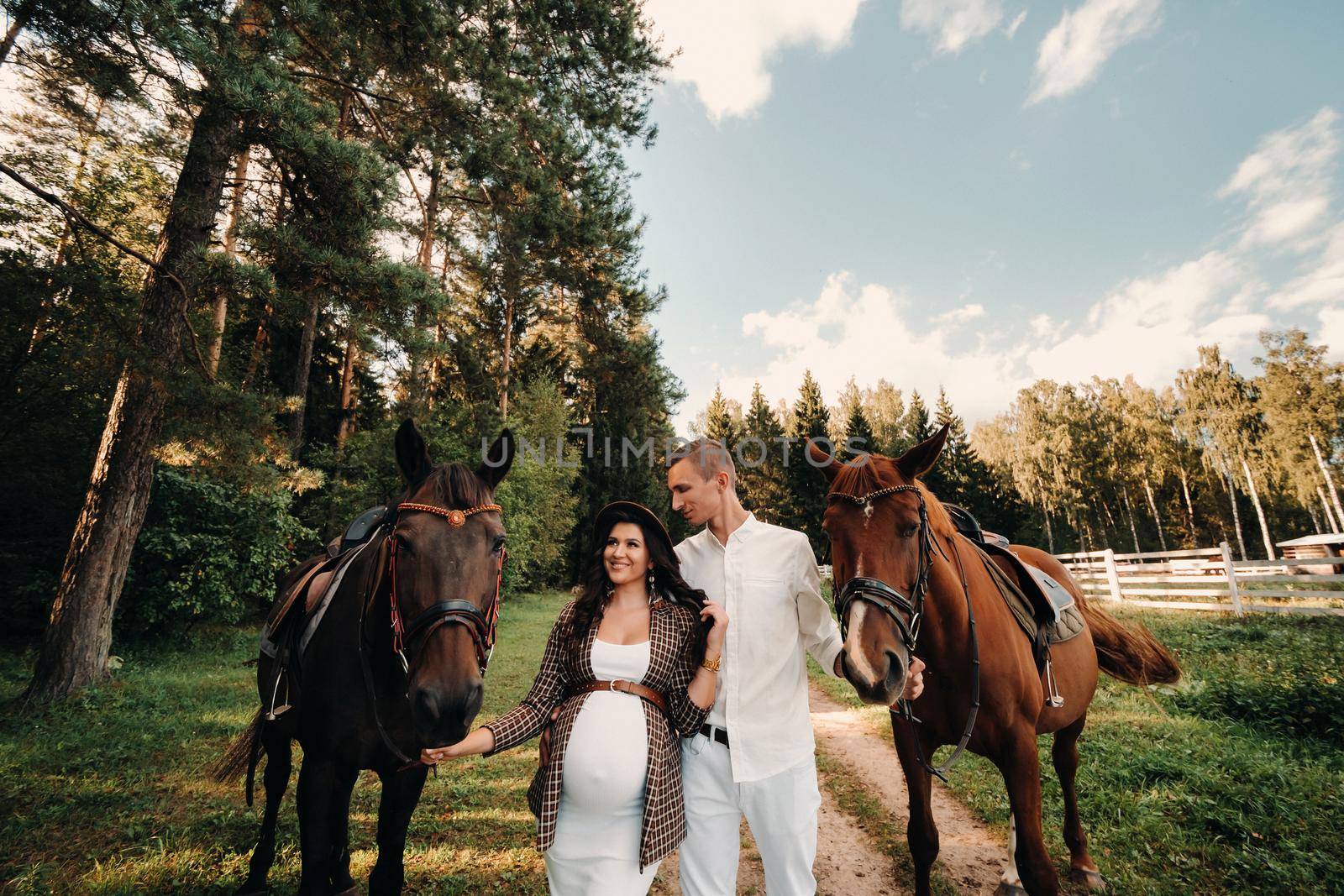 A pregnant woman in a hat with a man in white clothes walking with horses in nature. A family waiting for a child walks in the woods.