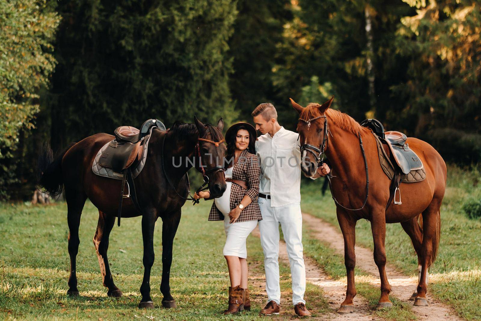 a pregnant girl in a hat and her husband in white clothes stand next to horses in the forest in nature.Stylish pregnant woman with a man with horses.Family. by Lobachad