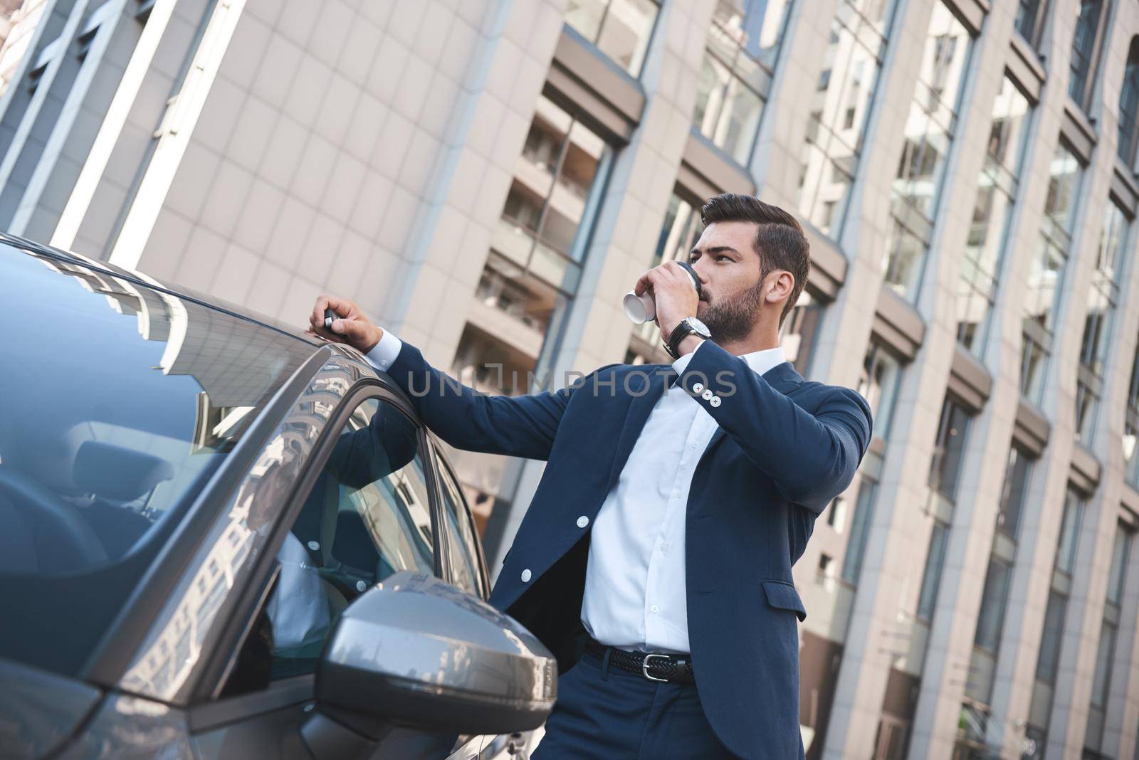 Confident businessman drinking coffee to go while standing near car on street