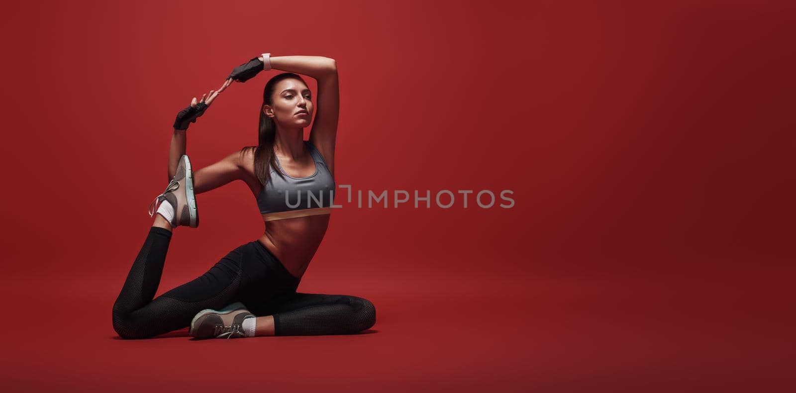 Stretching away the stress. Sportswoman sitting over red background, stretching her body by friendsstock