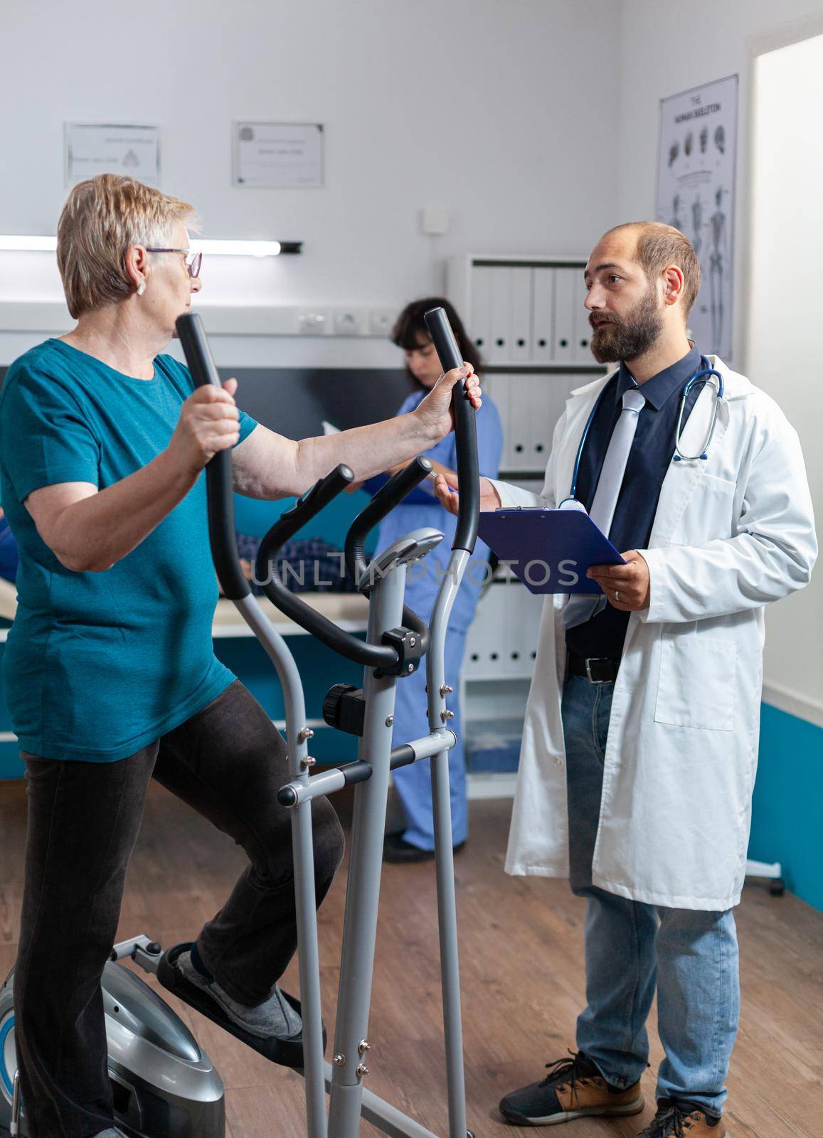 Doctor consulting elder patient doing gymnastics for recovery at physiotherapy clinic. Aged woman using stationary electrical bike for physical exercise and workout while talking to medic