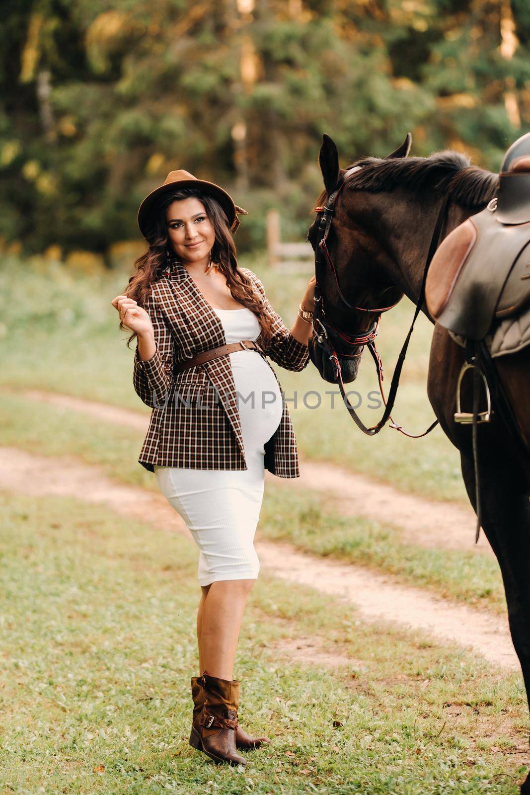 pregnant girl with a big belly in a hat next to horses in the forest in nature.Stylish pregnant woman in the brown dress with the horses