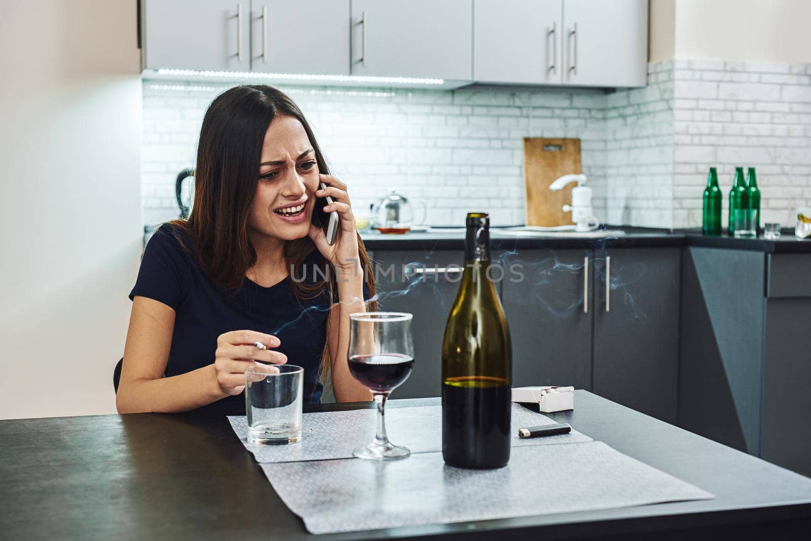 Portrait of young woman in despair, smoking cigarette while sitting at table, talking on the phone at home. Female alcoholism concept. Protest in the treatment of alcohol addiction.