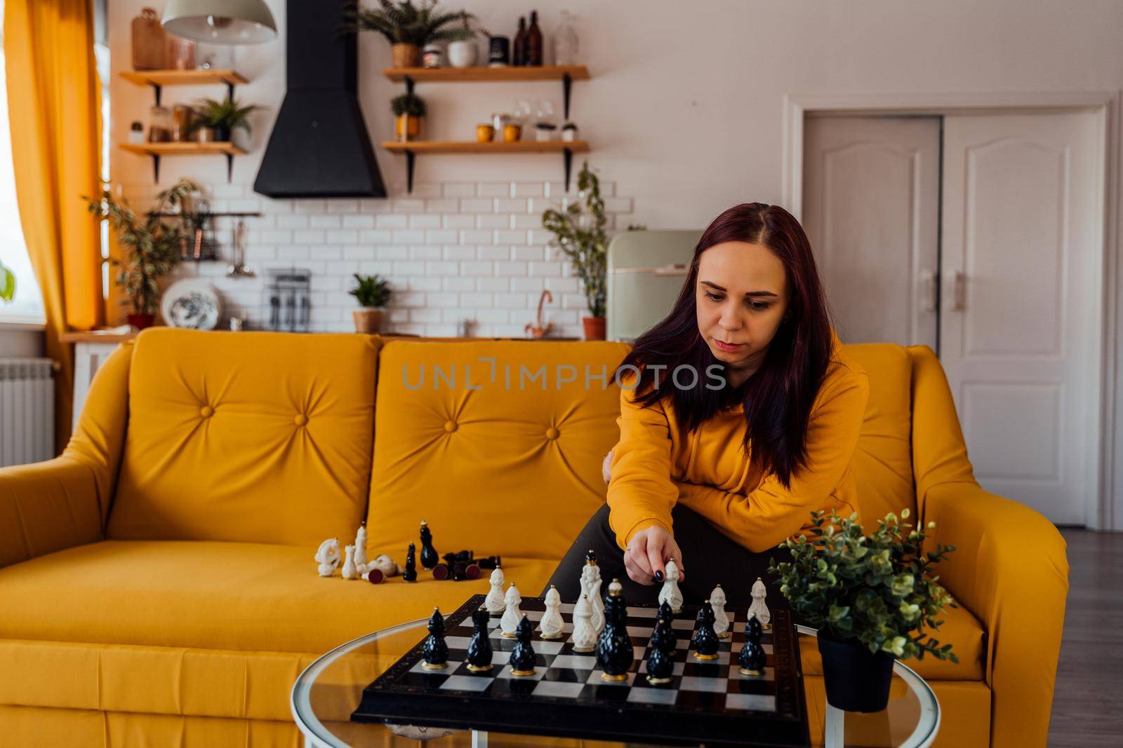 Young woman sitting on yellow sofa and playing chess in room. Female playing in logical board game with herself. by epidemiks