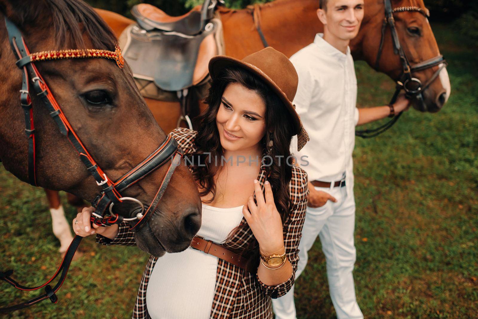 a pregnant girl in a hat and her husband in white clothes stand next to horses in the forest in nature.Stylish pregnant woman with a man with horses.Family