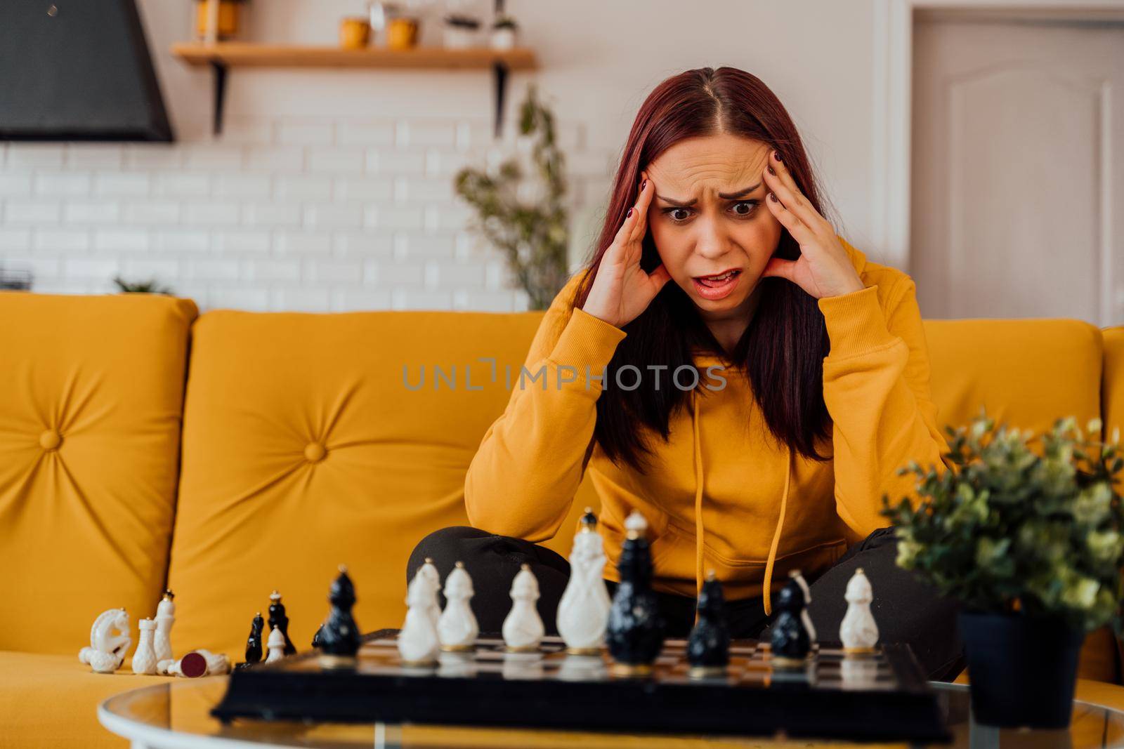 Young upset woman playing chess sitting on sofa. Distressed female plays in logical board game with herself in room