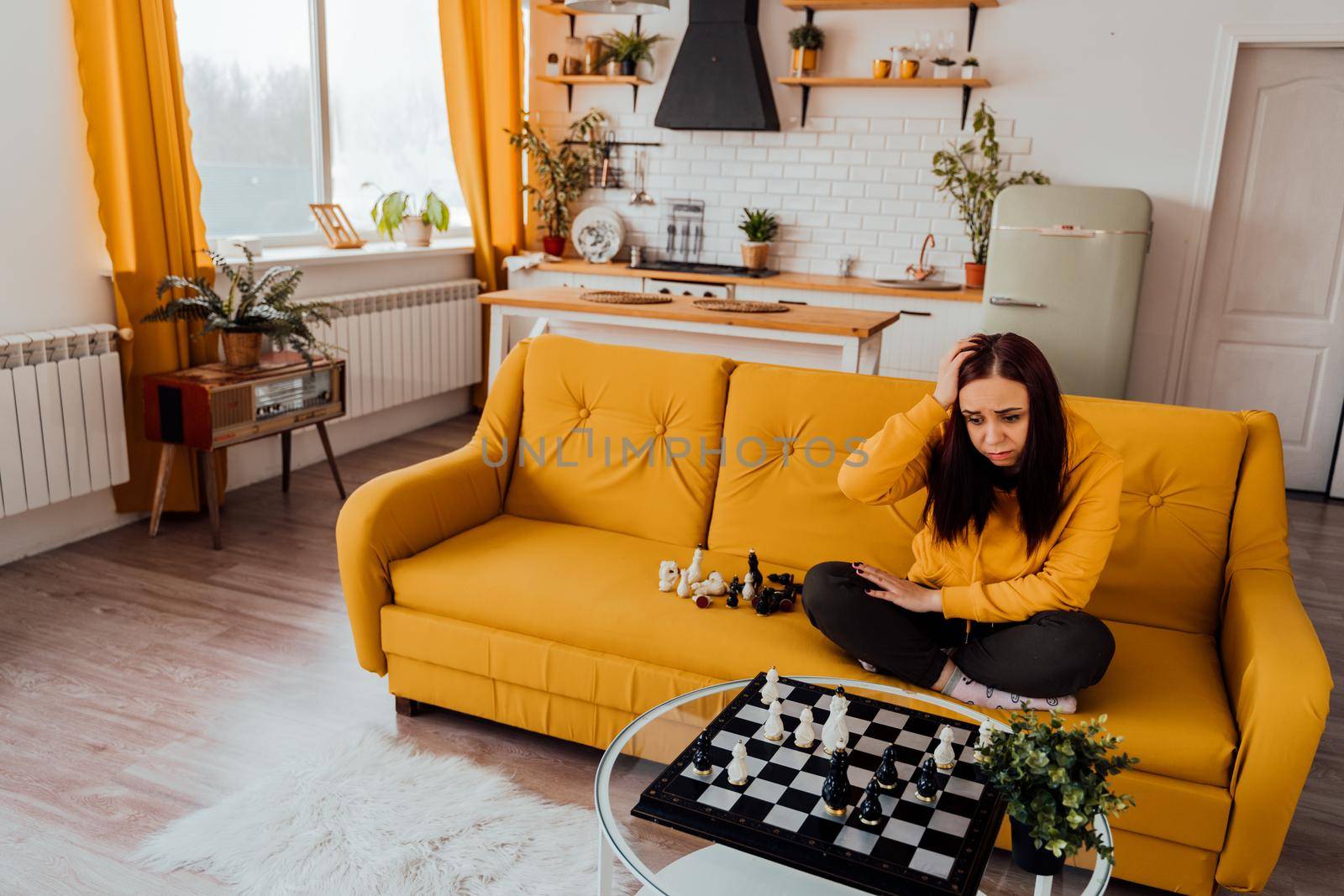 Young upset woman playing chess sitting on sofa. Distressed female plays in logical board game with herself in room
