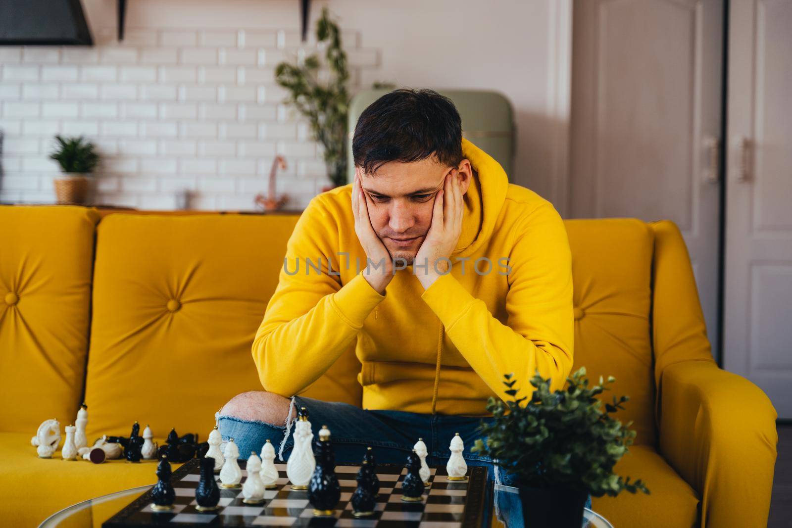 Young man sitting on yellow sofa and playing chess in room. Male playing in logical board game with himself. by epidemiks