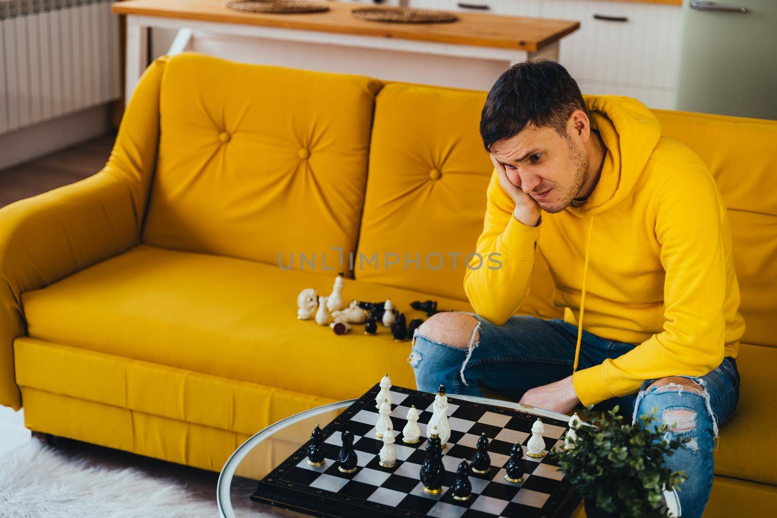 Young man sitting on yellow sofa and playing chess in room. Male playing in logical board game with himself. by epidemiks