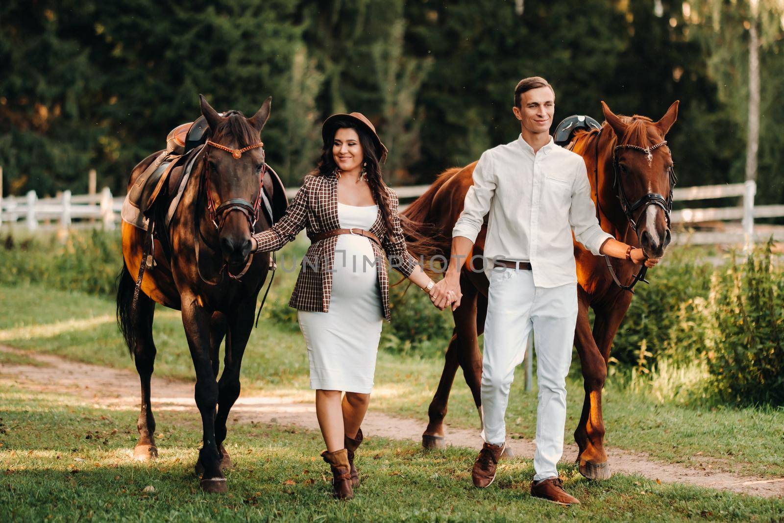 a pregnant girl in a hat and her husband in white clothes stand next to horses in the forest in nature.Stylish pregnant woman with a man with horses.Family