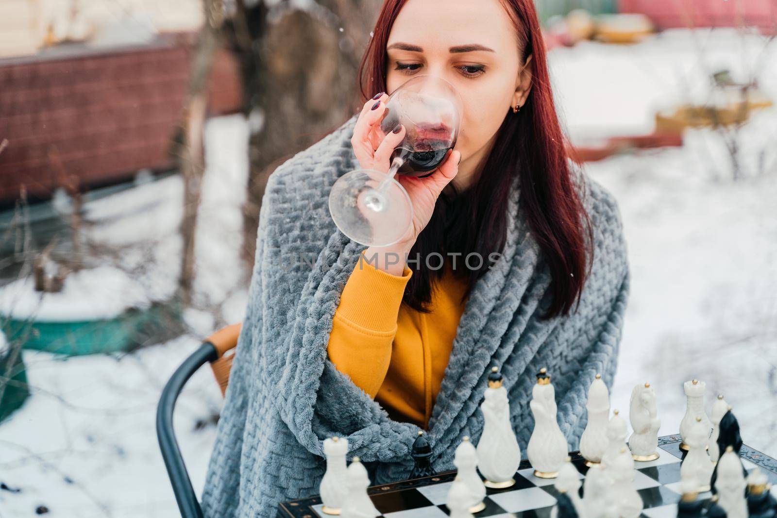 Young woman playing chess and drinking red wine in yard. Female wrapped in grey plaid with alcohol sitting on street playing in board game in winter season. by epidemiks