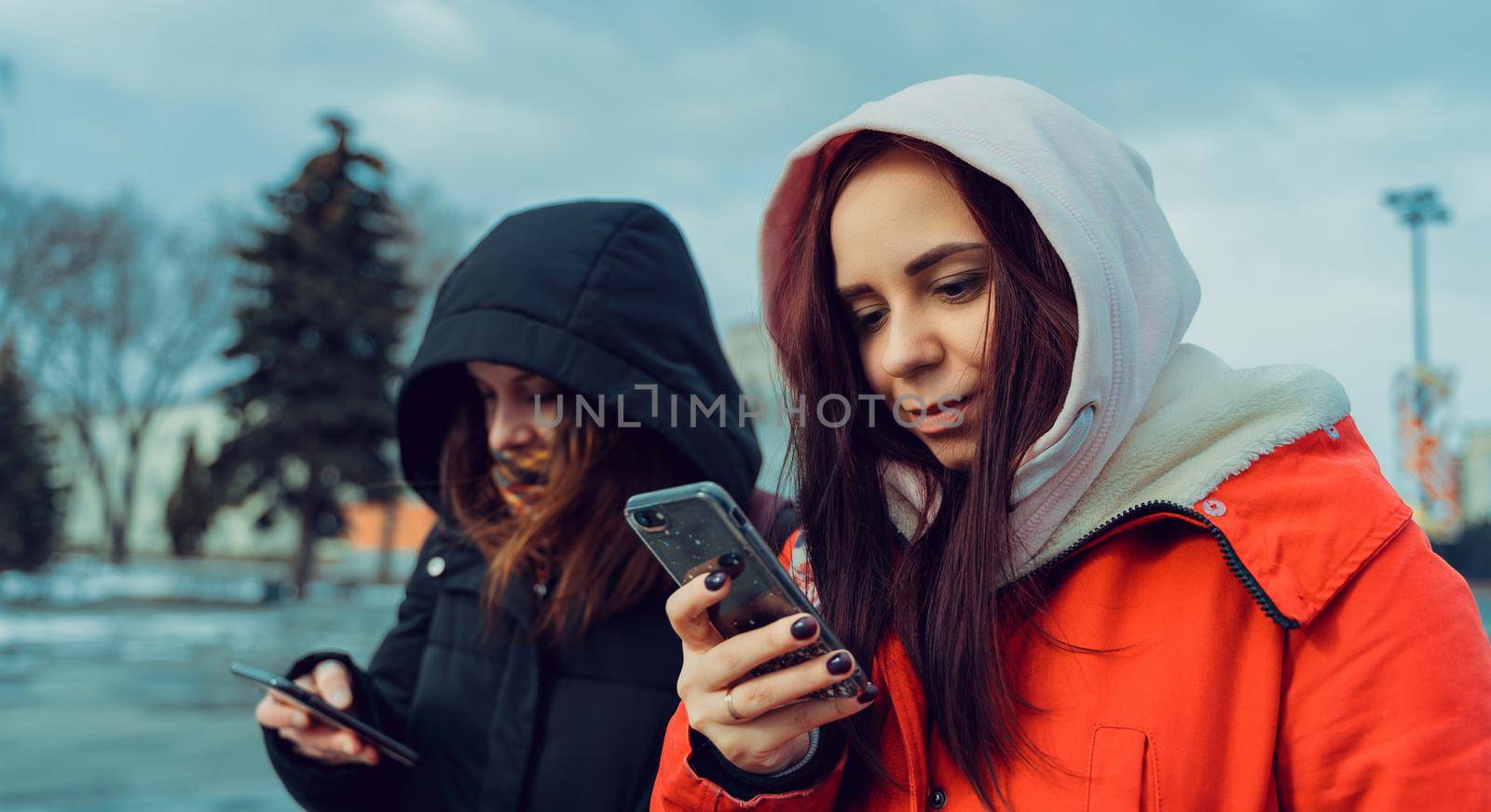 Close up of young women browsing smartphone in city park. Pretty females in hoods using mobile phone