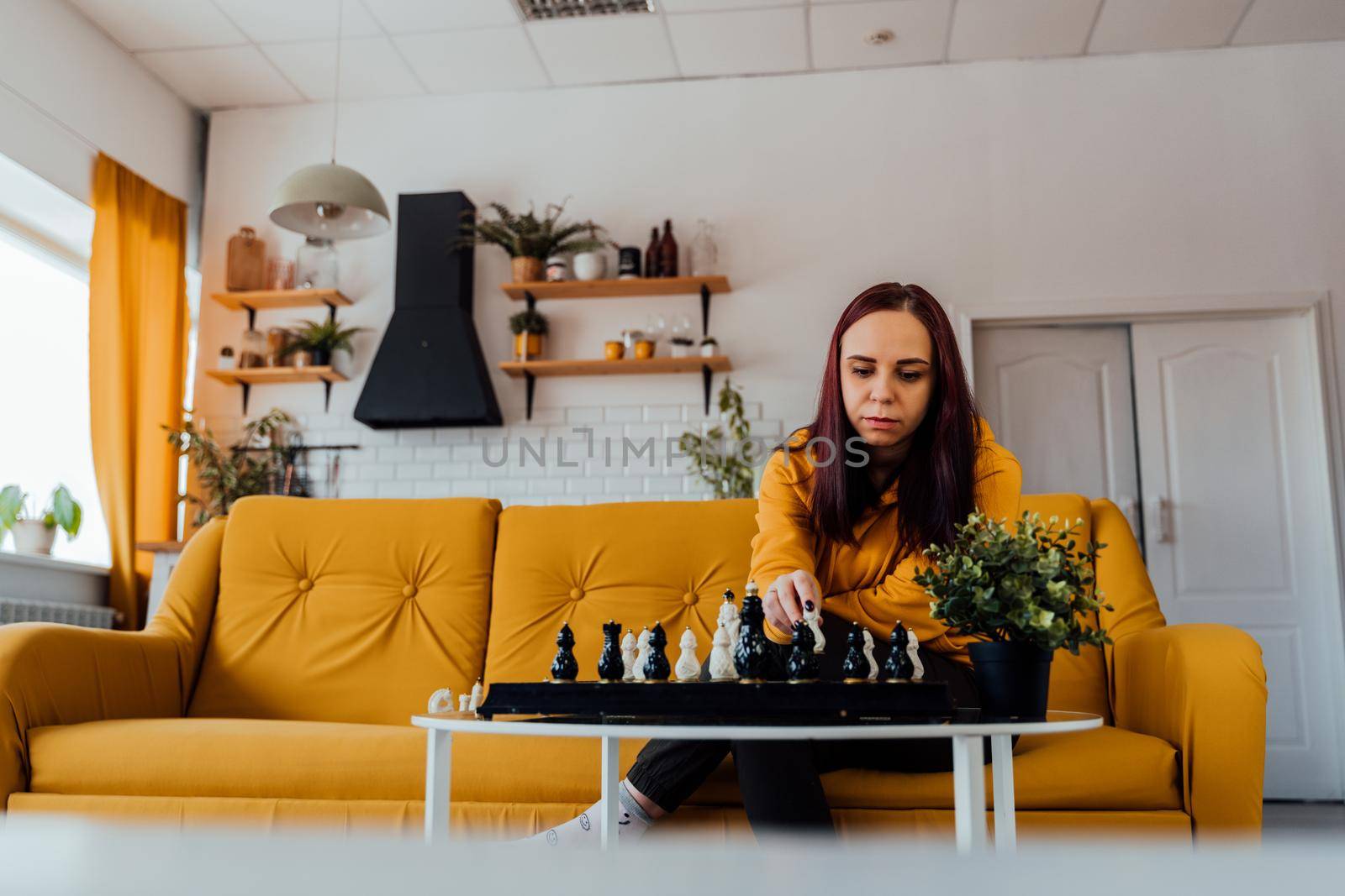 Young woman sitting on yellow sofa and playing chess in room. Female playing in logical board game with herself