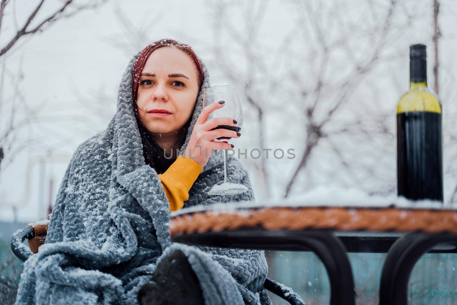 Young woman sitting in yard and drinking red wine in snowy weather. Female wrapped in grey plaid sitting on street with alcohol in winter season. by epidemiks