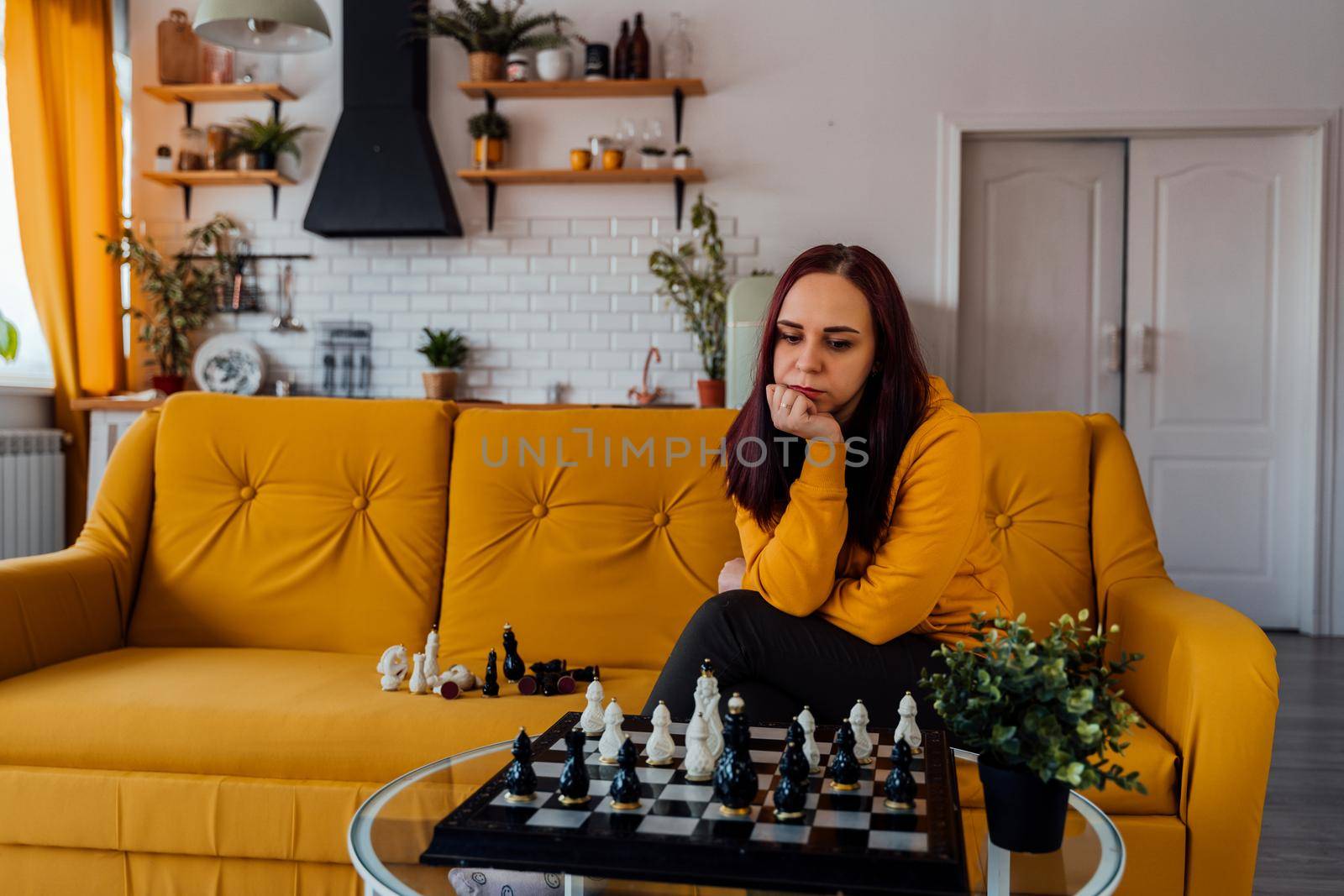 Young woman sitting on yellow sofa and playing chess in room. Female playing in logical board game with herself