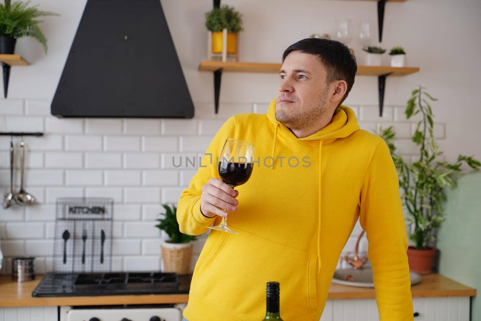 Relaxed young man poses with glass of red wine, standing at kitchen table. Adult guy resting with alcohol in kitchen