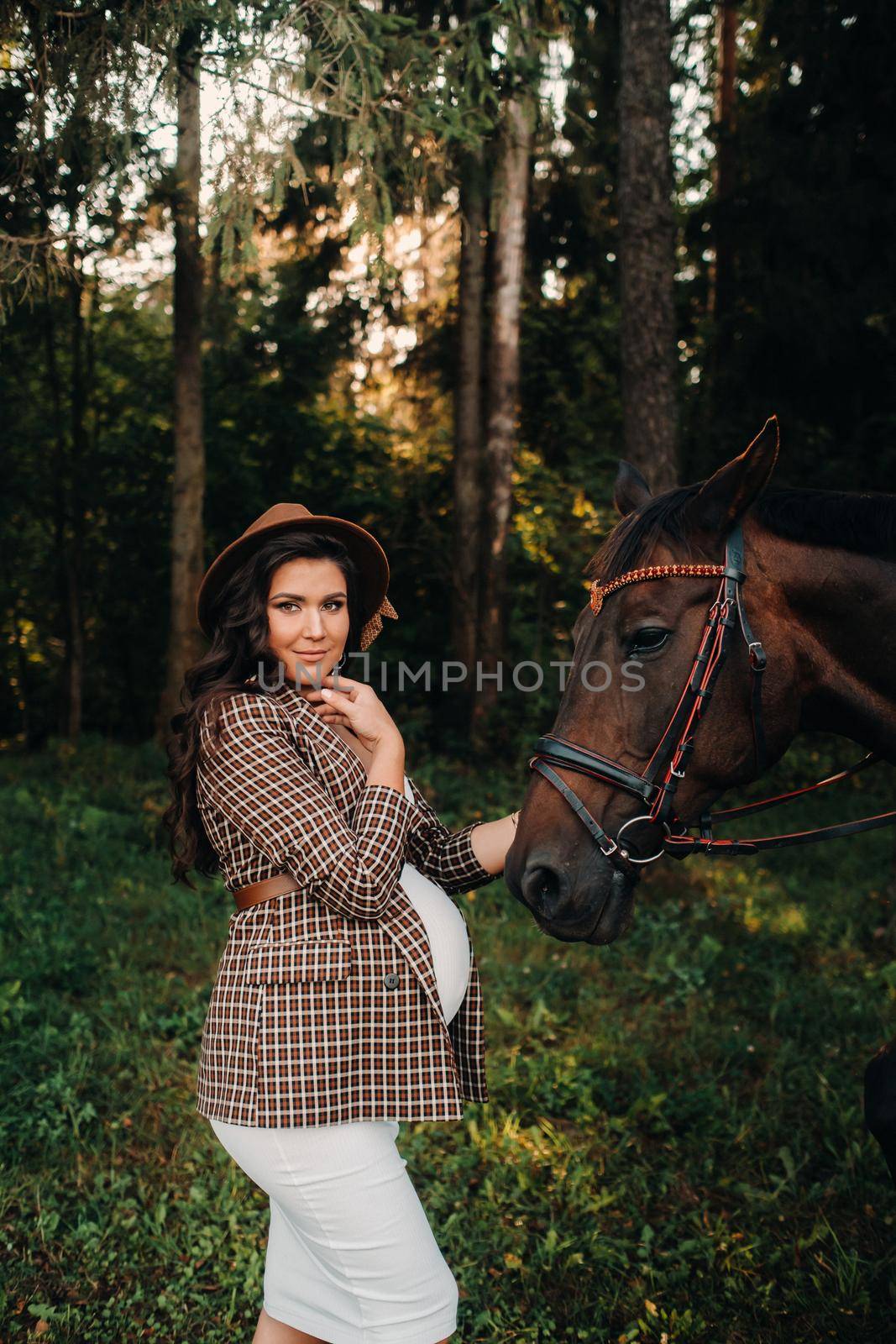 pregnant girl with a big belly in a hat next to horses in the forest in nature.Stylish girl in white clothes and a brown jacket