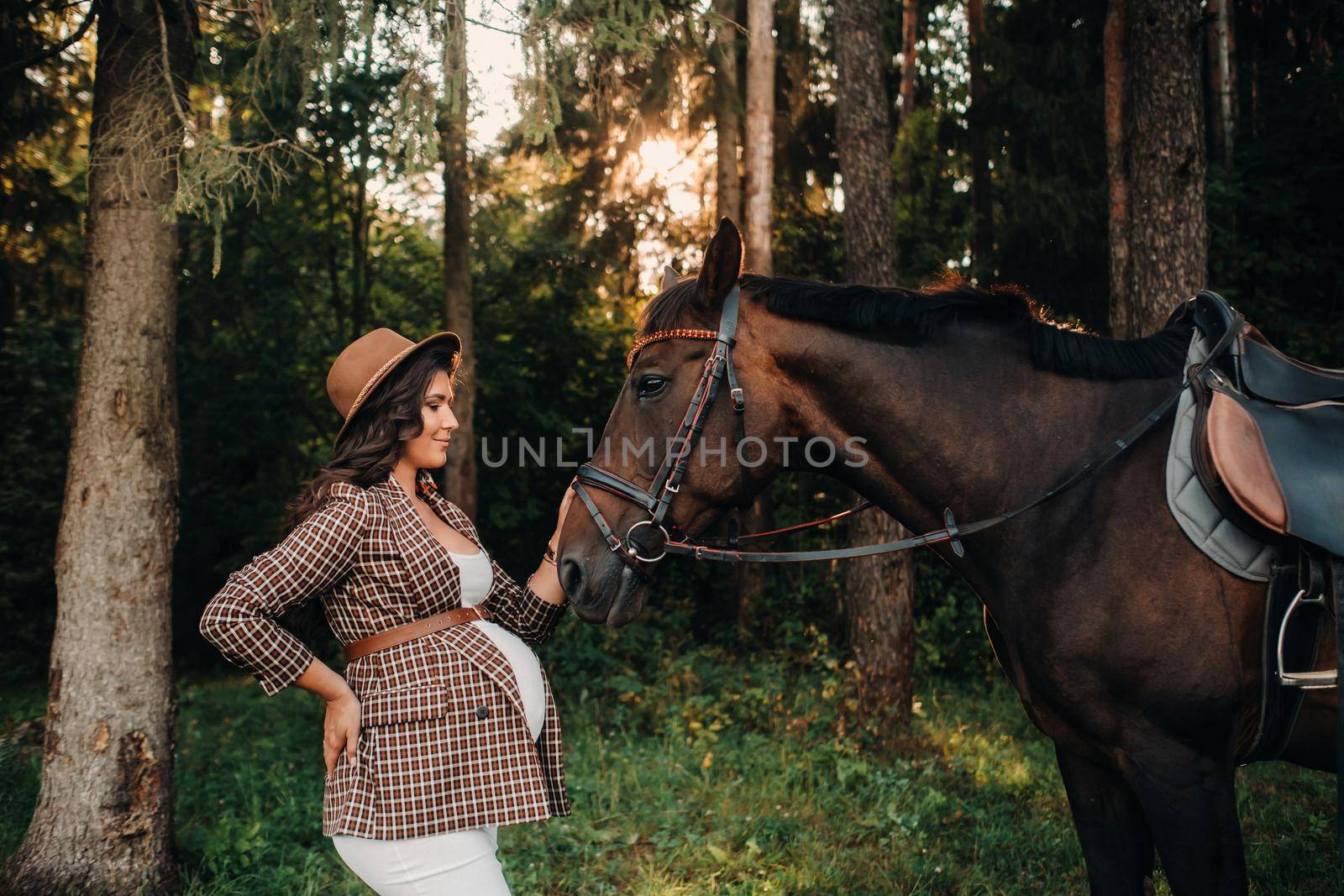 pregnant girl with a big belly in a hat next to horses in the forest in nature.A stylish pregnant woman in a white dress and brown jacket with horses. by Lobachad