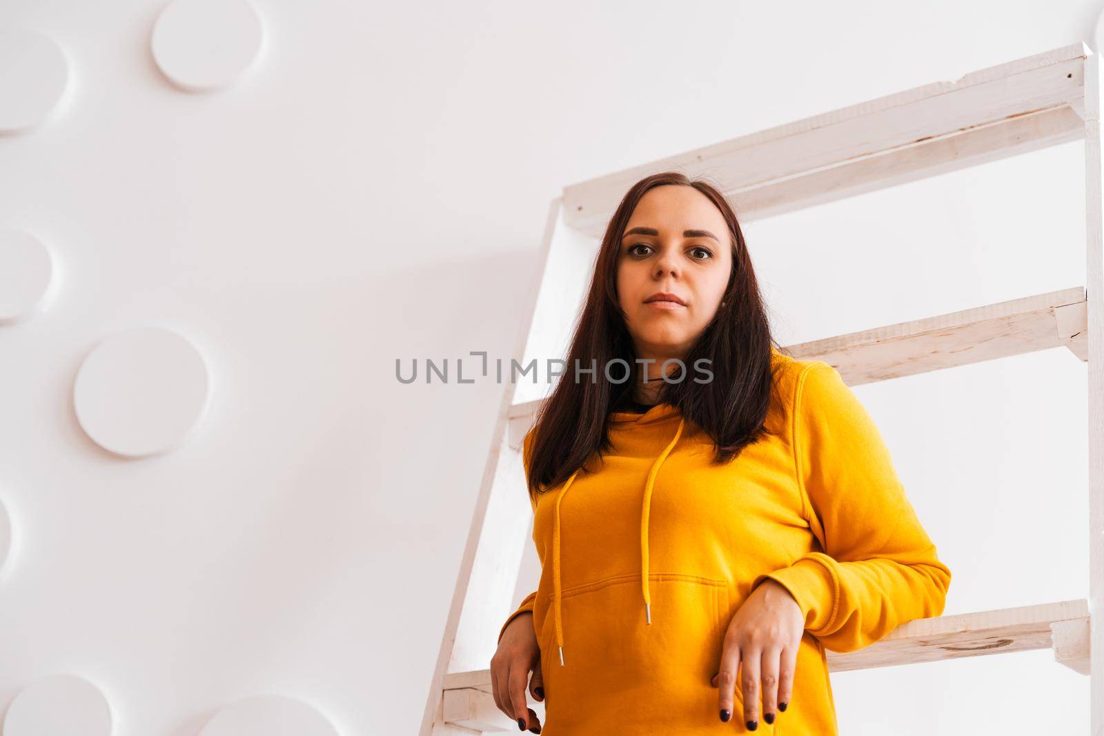 Portrait of young woman posing on white background. Pretty brunette in yellow hoodie near stepladder on background of white wall