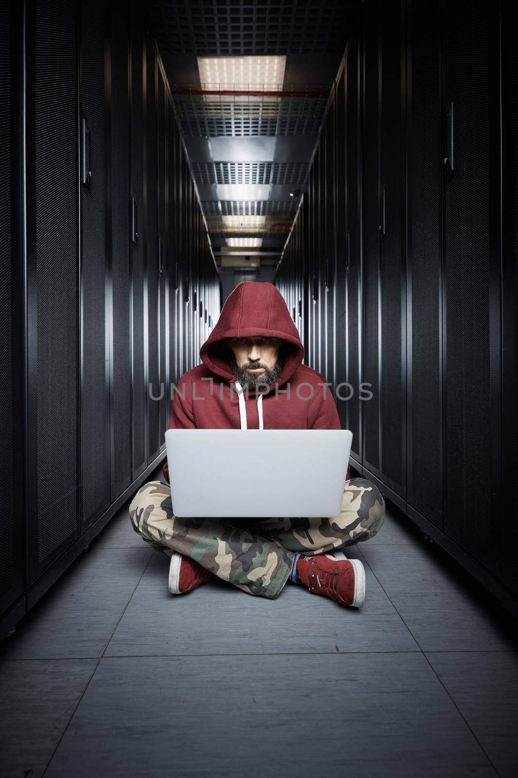 The hacker in a hood sits with the computer among servers, the burglar. Storage of data, cloudy services, system breaking, attack of hackers by vladimirdrozdin