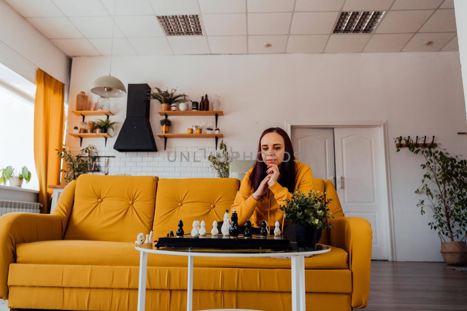 Young woman sitting on yellow sofa and playing chess in room. Female playing in logical board game with herself