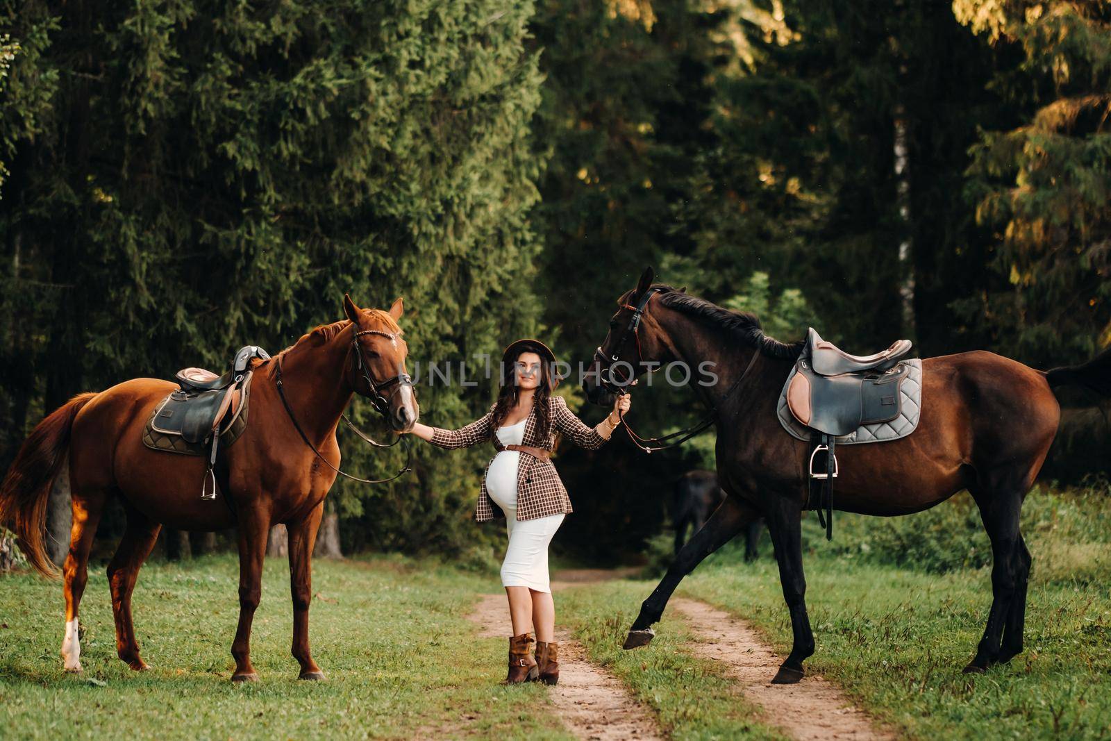 pregnant girl with a big belly in a hat next to horses in the forest in nature.Stylish girl in white clothes and a brown jacket