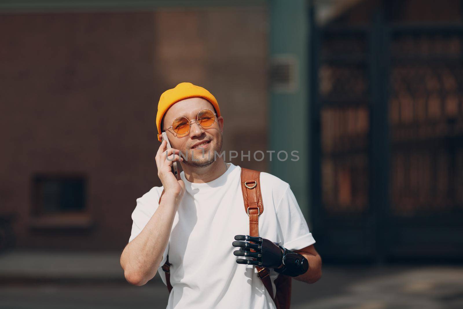 Young disabled man hold mobile phone artificial prosthetic hand in casual clothes and backpack walking at sunny city street outdoor