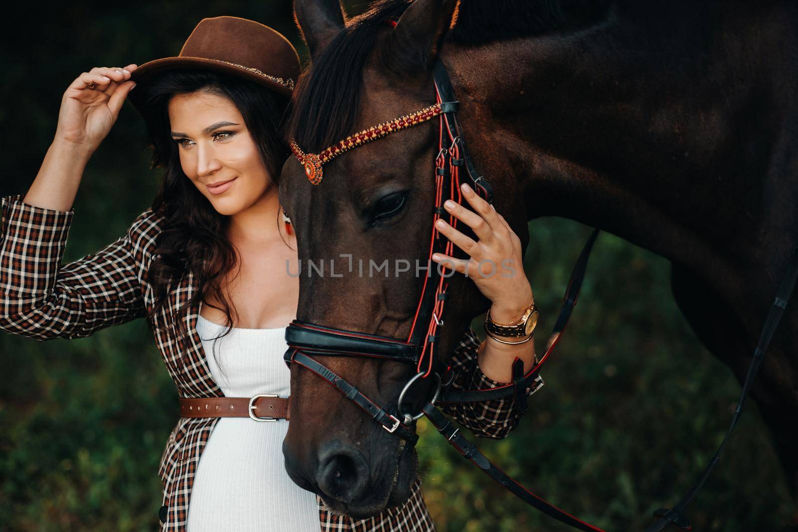 pregnant girl with a big belly in a hat next to horses in the forest in nature.Stylish girl in white clothes and a brown jacket