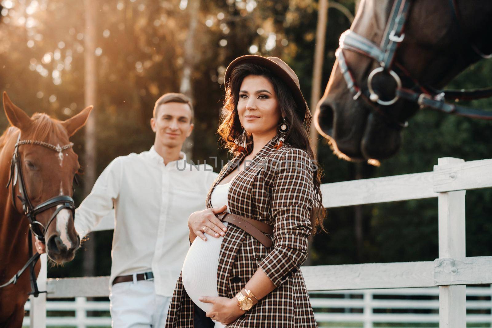 a pregnant girl in a hat and a man in white clothes stand next to horses near a white fence.Stylish pregnant woman with a man with horses.Married couple