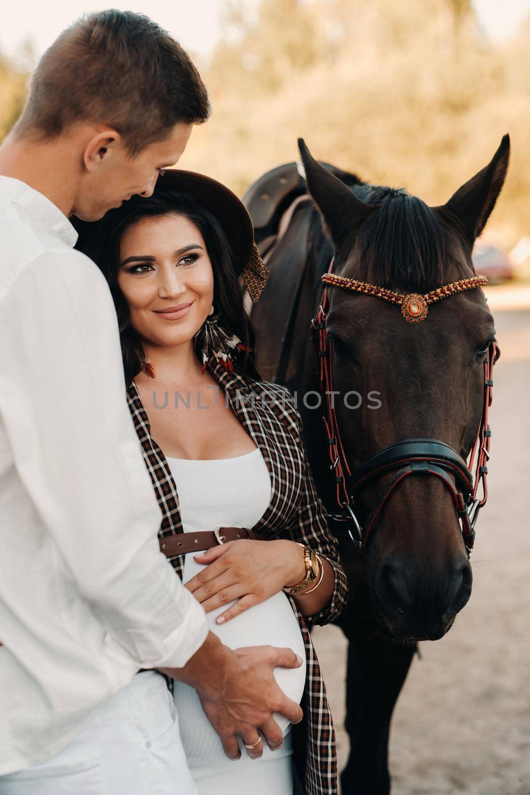 a pregnant girl in a hat and a man in white clothes stand next to horses near a white fence.Stylish pregnant woman with a man with horses.Married couple