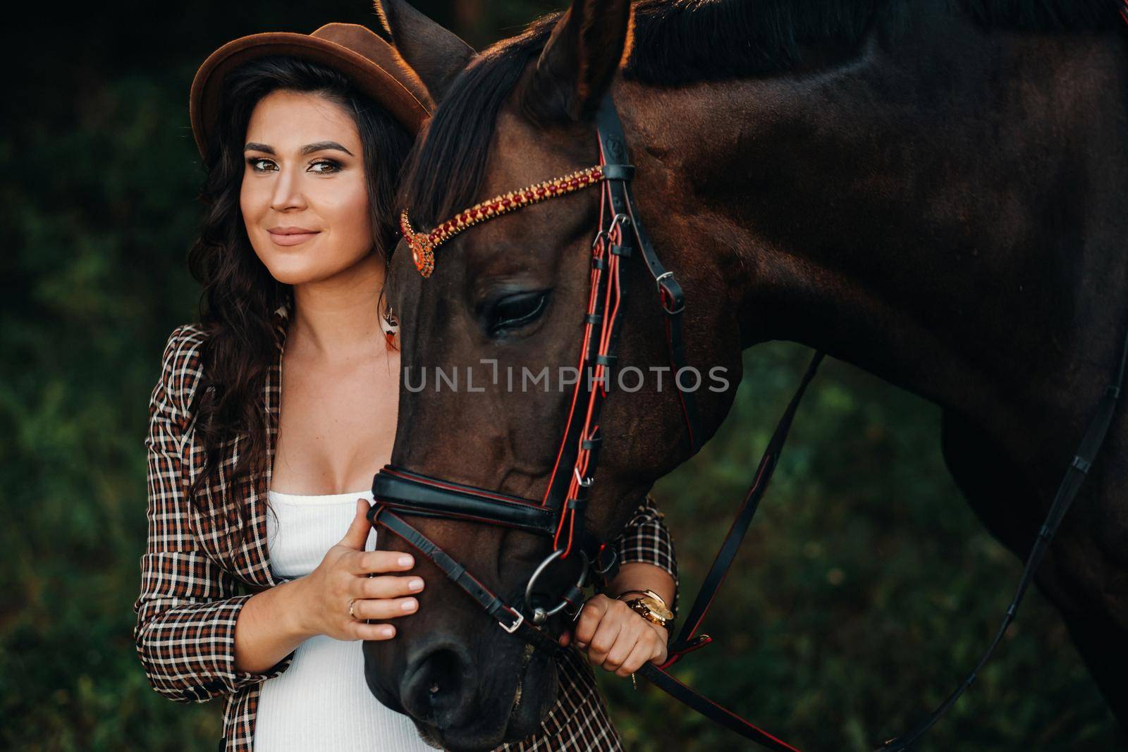 pregnant girl with a big belly in a hat next to horses in the forest in nature.Stylish girl in white clothes and a brown jacket