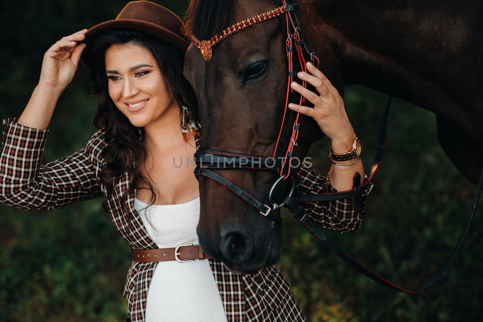 pregnant girl with a big belly in a hat next to horses in the forest in nature.Stylish girl in white clothes and a brown jacket