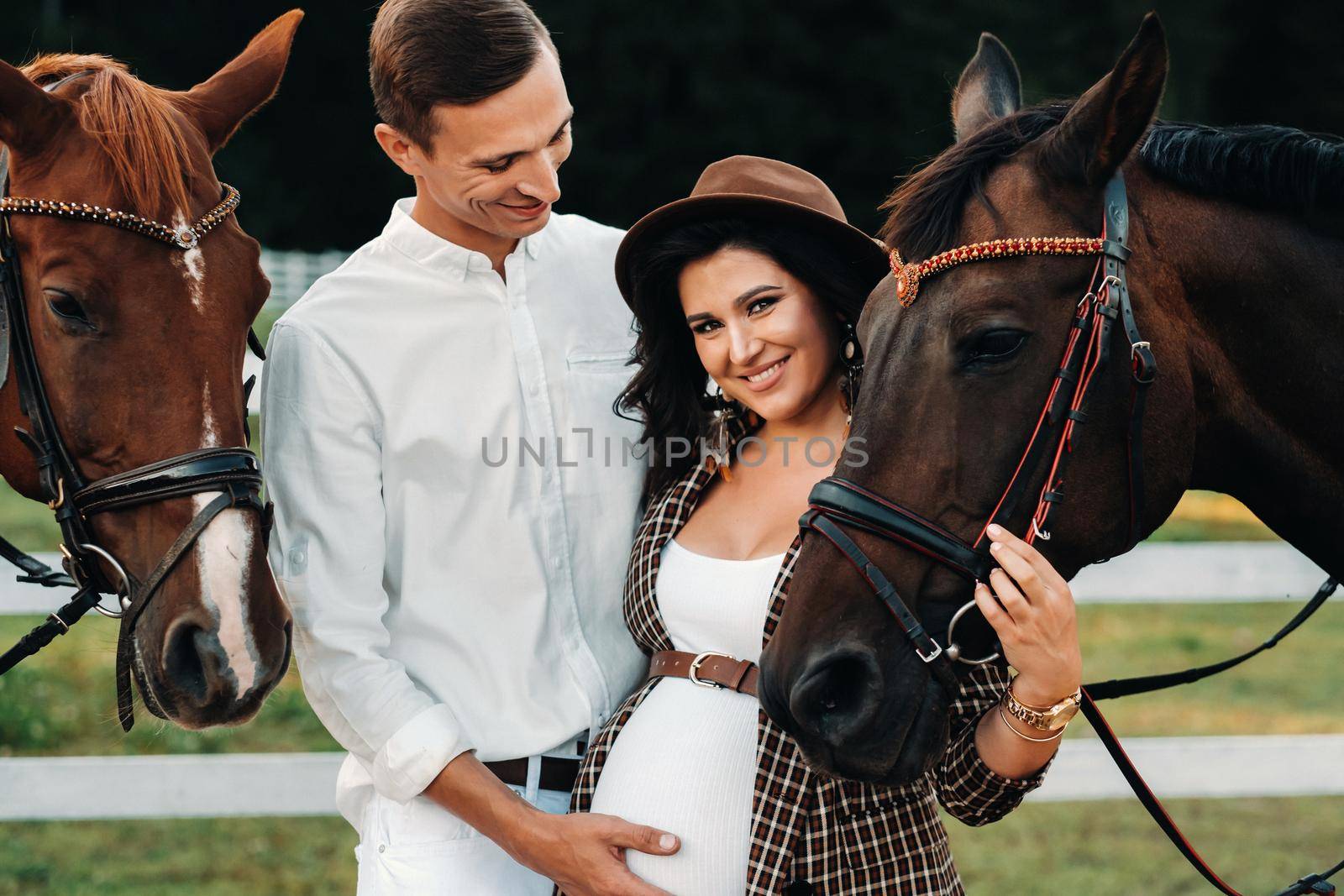 a pregnant girl in a hat and a man in white clothes stand next to horses at a White fence.Stylish family waiting for a child strolling in nature.