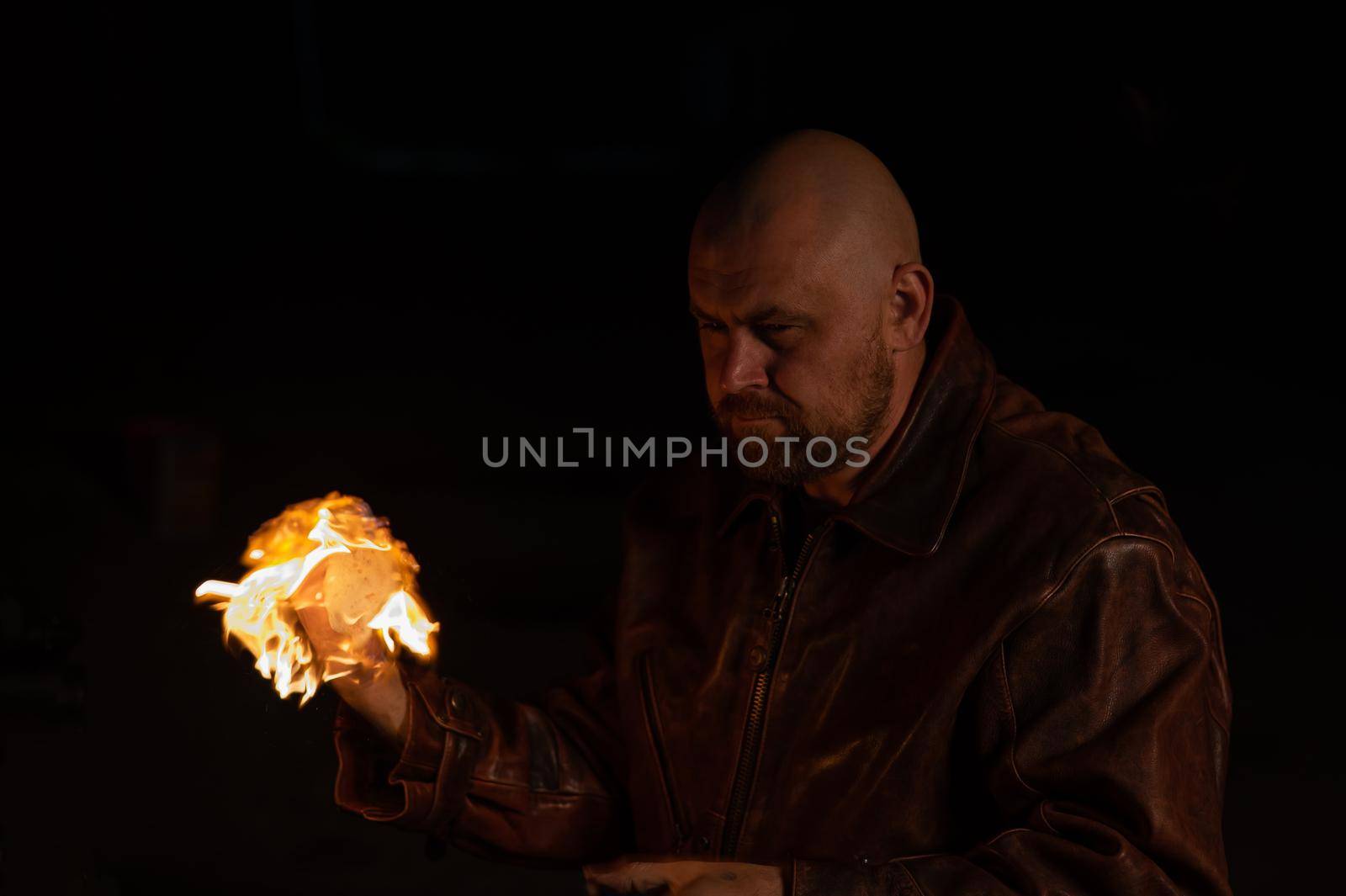 A bald man in a leather jacket holds a burning ball in his hand