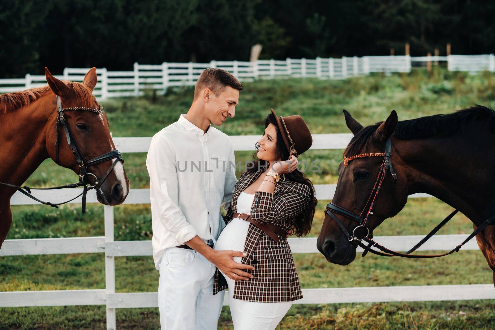 a pregnant girl in a hat and a man in white clothes stand next to horses near a white fence.Stylish pregnant woman with a man with horses.Married couple. by Lobachad