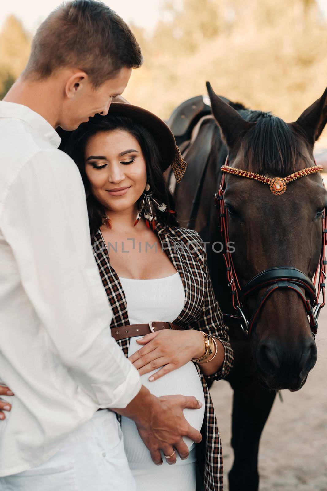 a pregnant girl in a hat and her husband in white clothes stand next to horses in the forest in nature.Stylish pregnant woman with a man with horses.Family. by Lobachad