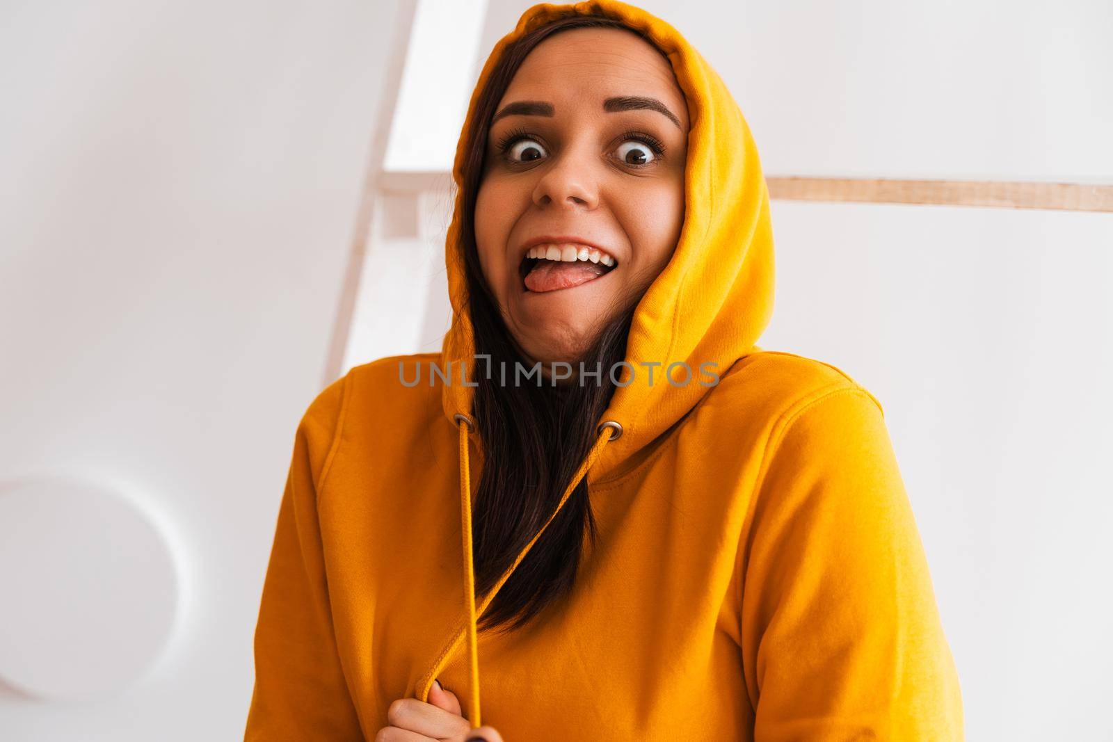 Portrait of young woman posing on white background. Pretty brunette in yellow hoodie near stepladder on background of white wall. by epidemiks